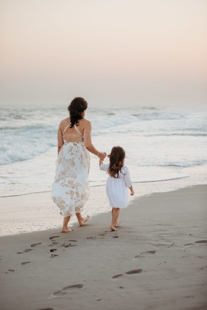 Mom and daughter walk holding hands on the beach at sunset.