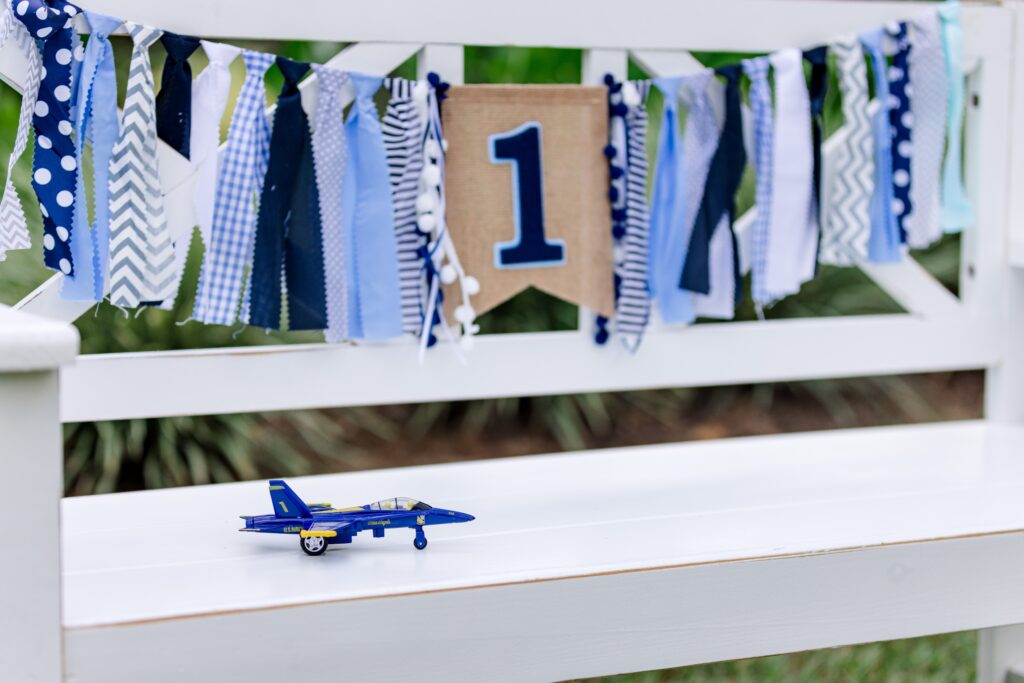 Navy Blue Angels - Baby's First Birthday Prop die cast toy jet sitting on white bench with blue birthday banner.