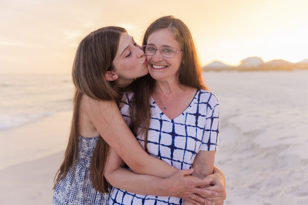 Teenage daughter kissing mom on the cheek during family photo session on the beach with a beautiful sunset behind them. 